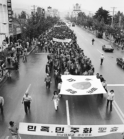 흑백사진입니다. 맨 앞엔 민족 민주화 성회, 전남대학교 총학생회 라고 써있는 현수막을 든 사람들과 그 뒤를 커다란 태극기를 여성 여덟명이 나누어 들고 걷고 있습니다. 그 뒤에는 많은 사람들이 양복을 입고 행진 중인 모습입니다. 